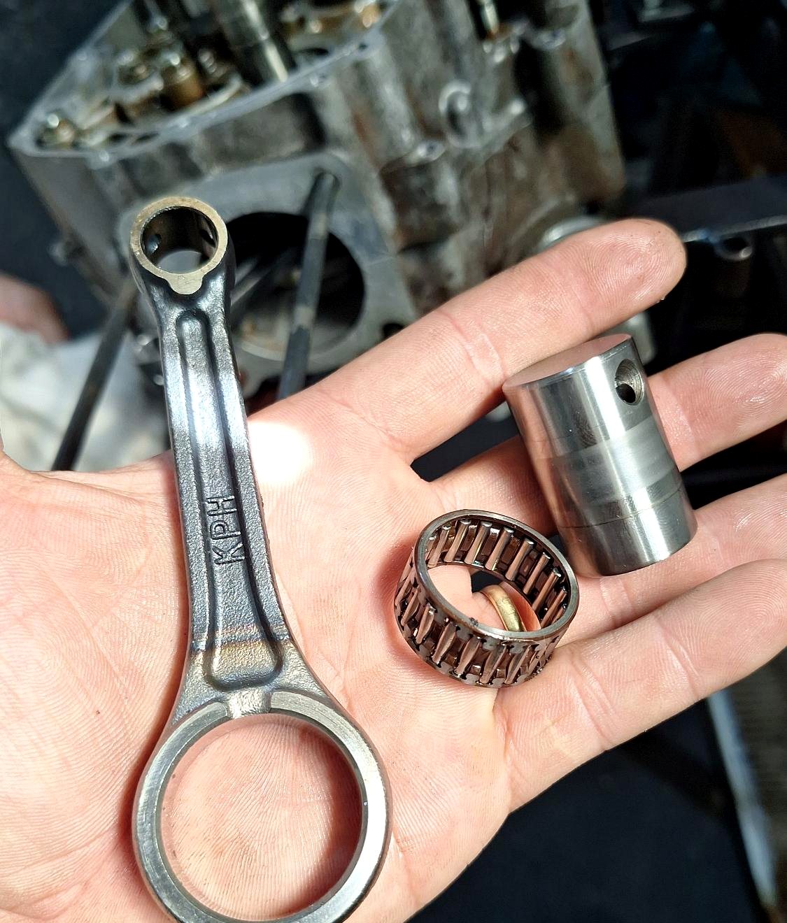 Mão segurando biela, rolamento de agulha e pino, com o motor aberto ao fundo.